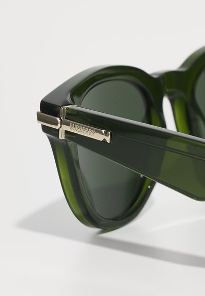 Zaļas acetāta saulesbrilles ar biezām rāmī, izmantojot zelta toņa dekoratīvo akcentu ar iegravētu "BURBERRY" un tumšiem stikliem.