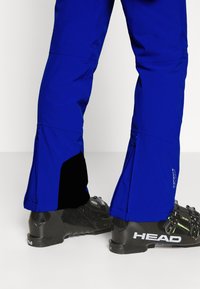 Jambes inférieures dans un pantalon de ski bleu vif avec des poignets renforcés en noir sur des bottes de ski HEAD noires, sur fond blanc.