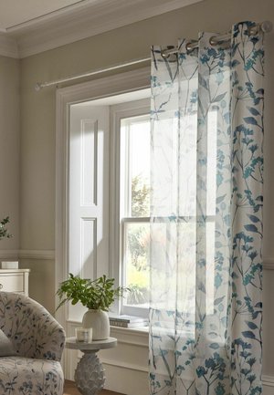 Durchscheinende weiße Vorhänge mit blauem Blumenmuster hängen an einem sonnenbeschienenen Fenster neben einem gemusterten Sessel und einem kleinen runden Tisch mit einer Pflanze.