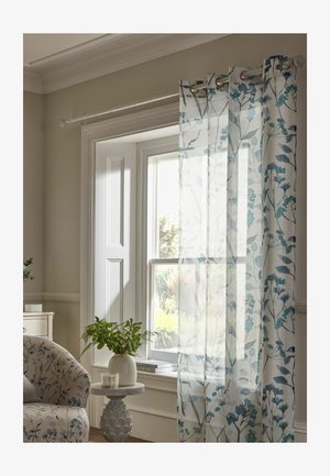 Durchscheinende weiße Vorhänge mit blauem Blumenmuster hängen an einem sonnenbeschienenen Fenster neben einem gemusterten Sessel und einem kleinen runden Tisch mit einer Pflanze.