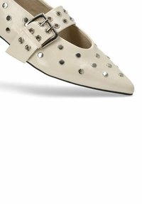 Zapato de punta en cuero crema con tachuelas redondas metálicas y una amplia correa de hebilla en la parte media del pie. Suela negra.