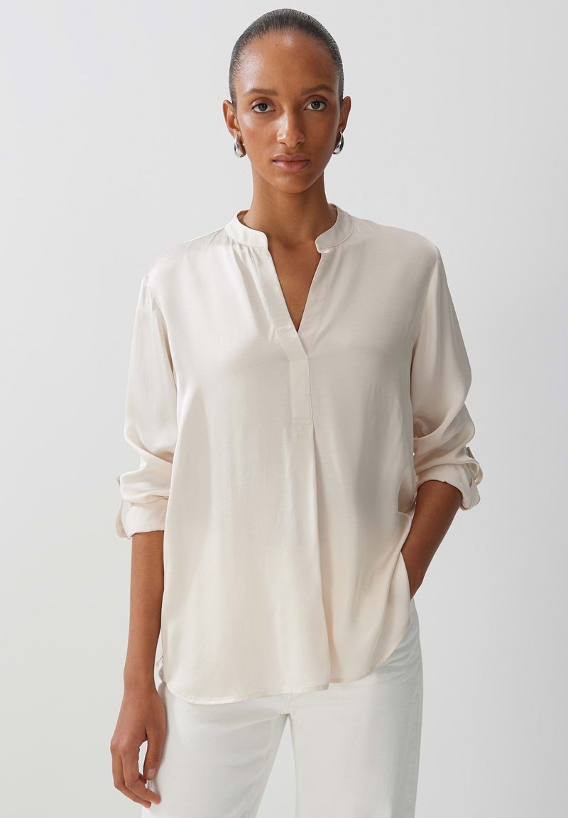 Blusa lunga di colore beige chiaro, con colletto alla mandarino, texture morbida, maniche a tre quarti arrotolate e vestibilità comoda.