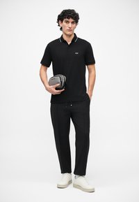Homem vestido com uma camisa polo preta e calças pretas, segurando uma bolsa cinza com ziper, de pé com uma mão no bolso e ténis brancos.