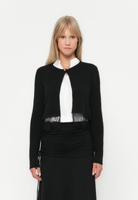 Crna skraćena cardigan jakna s okruglim izrezom i čipkastim detaljima preko bijele bluze. Cardigan ima mali zatvarač na dugme na vratu.