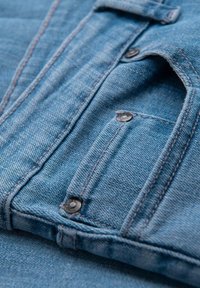 Detailní záběr modré džínsoviny s malou kapsičkou na mince, viditelným šitím a kovovými nýty.