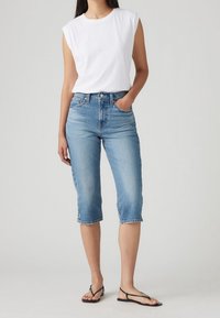 Bílý bezrukávový bavlněný top spárovaný se světle modrými zkrácenými džíny, které mají pět kapes a roztrhané lemy. Černé ploché sandály dokončují vzhled.