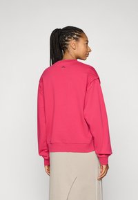 Sweatshirt fúcsia com mangas compridas, punhos e barra canelados, decote redondo, com um pequeno detalhe de logótipo na parte de trás. Textura de tecido suave.