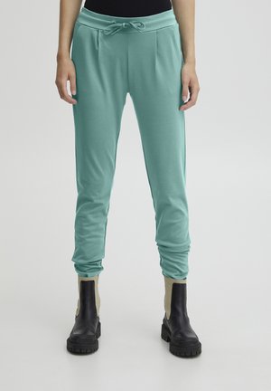 Kvinde iført grønblå joggers med snøre i taljen og sorte chunky ankelstøvler på enkel lys baggrund.