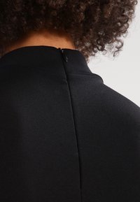 Czarna sukienka z wysokim dekoltem, z tylnym zamkiem błyskawicznym i gładkim, teksturowanym materiałem. Projekt jest elegancki i dopasowany.