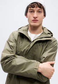 Wasserfeste Jacke in Olivgrün mit einer Reißverschlusstasche auf der Brust, einer verstellbaren Kapuze und elastischen Bündchen. An einem Modell mit grauer Mütze präsentiert.