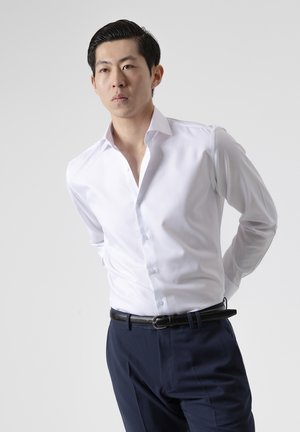 Hombre asiático que lleva una camisa blanca y pantalones azul marino, de pie con las manos detrás de la espalda contra un fondo liso.