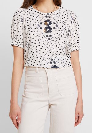 Vrouw met een witte blouse met zwart bloem- en stippenpatroon, pofmouwen, gecombineerd met witte broek met hoge taille en gelaagde gouden kettingen.