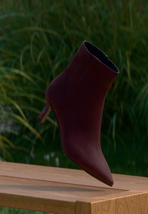 Stivaletto viola scuro con tacco alto che fluttua sopra una superficie di legno con fogliame verde sullo sfondo.