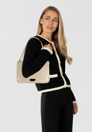 Borsa a tracolla in pelle crema con catena dorata, forma rettangolare e dettaglio della chiusura. Il modello indossa un cardigan nero con dettagli crema.