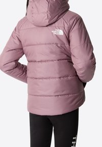 Osoba w fioletowej kurtce puchowej The North Face z kapturem, z rękami w kieszeniach, w połączeniu z czarnymi spodniami z białym napisem na udzie.