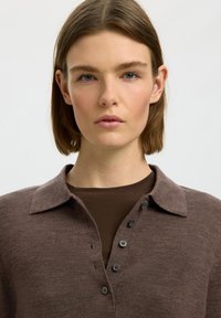 Ung person med glat brunt hår iført en brun knappet sweater over en mørkebrun skjorte mod en ensfarvet baggrund.