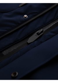 Detail zblízka na černý zip navy modré bundy a síťovou podšívku vedle kovového patentku.
