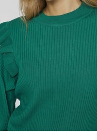 Långärmad grön ribbad tröja med volangdesign på axlarna. Tyget verkar mjukt med en texturerad yta.