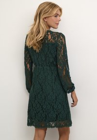 Robe en dentelle vert foncé avec des manches longues, motif floral, taille cintrée et ourlet festonné. La texture transparente permet d'apercevoir la peau en dessous.