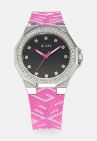 Guess-klocka med svart urtavla, silverfärgad bezel prydd med kristaller och rosa mönstrad silikonrem. Har rosa visare och fyrkantiga timmarkörer.