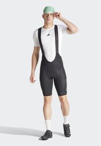 Salopette da ciclismo nera con bretelle bianche, abbinata a una maglietta bianca aderente e un cappellino verde chiaro. Caratteristiche includono un sedile imbottito e dettagli riflettenti.