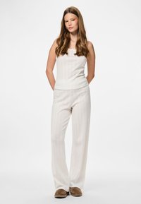 Lengvas, tekstūruotas dviejų dalių kostiumas, sudarytas iš rankovių neturinčio viršaus ir plačių kojų kelnių švelnaus kreminio atspalvio, derinamas su rudos spalvos šlepetėmis.