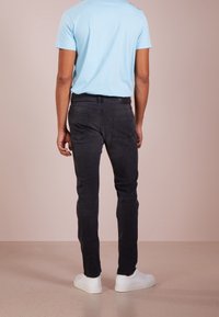 Muž v světle modrém tričku, černých džínách a bílých teniskách stojící odvrácený proti béžovému pozadí.