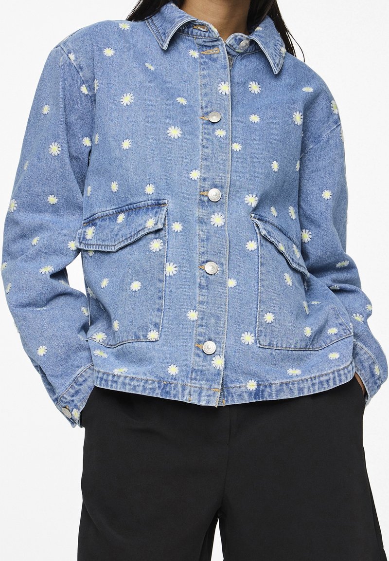 Vaaleansininen farkkutakki, jossa on valkoista päivänkakkaraembroidausta, nappikiinnitys, kaksi etutaskua ja klassinen kaulustyyli.