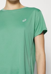 Camiseta deportiva de manga corta en tejido verde claro, con cuello redondo y un pequeño logo reflectante en el pecho.