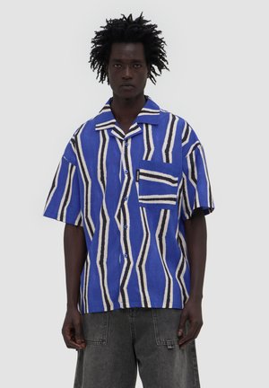 Mężczyzna z kręconymi włosami, ubrany w niebieską, białą i czarną geometrycznie paskowaną koszulę z krótkim rękawem oraz ciemnoszare dżinsy na jednolitym tle.