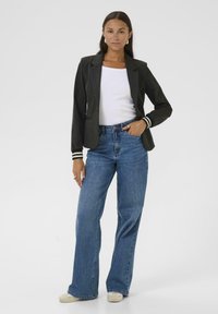 Blazer gris foncé avec des poignets rayés blancs, associé à un jean large taille haute bleu clair et un haut blanc. Modèle se tenant avec les mains sur les hanches.