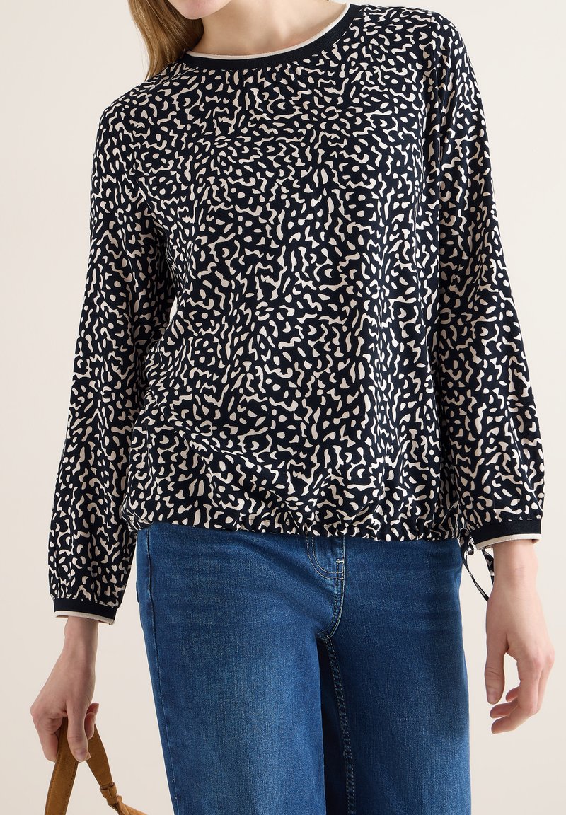 Femme portant un chemisier à manches longues à motifs bleu marine et beige avec un ourlet élastique, ainsi qu'un jean bleu, tenant une sangle marron dans sa main gauche.