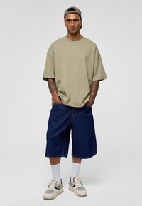 Khaki oversized t-shirt met korte mouwen, gecombineerd met donkere denimshorts. Witte crew sokken en multicolore sneakers maken de outfit compleet.