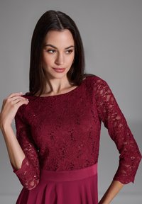 Robe bordeaux avec des manches longues en dentelle et des détails en sequins, affichant un col rond et une taille cintrée qui se prolonge par une jupe fluide.