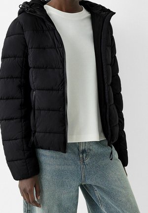 Osoba w czarnej pikowanej kurtce z kapturem, na białej koszuli i jasnoniebieskich dżinsach, na jednolitym tle.