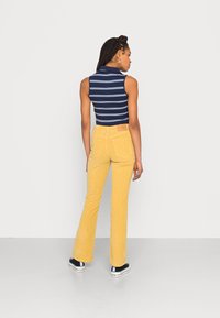 Top às riscas em azul-marinho e branco sem mangas combinado com calças em veludo cotelê amarelo mostarda em corte flare, apresentando um rótulo com um patch na parte de trás.
