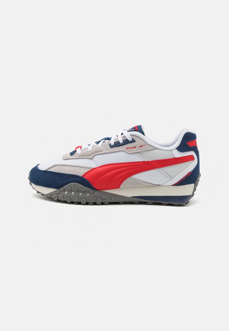 Baskets Puma blanches, grises, bleu marine et rouges avec une bande latérale rouge, fermeture à lacets et semelle texturée, présentées en profil latéral sur fond blanc.