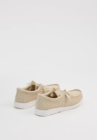 Zapatillas de malla beige con suelas y cordones blancos, que presentan una superficie texturizada y forma redondeada. Diseño simple para uso casual.