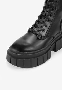 Stivale in pelle nera con lacci, suola piatta spessa e scanalata e dettaglio metallico sui lacci su sfondo bianco.