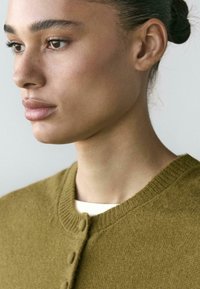 Cardigan in maglia verde oliva con collo rotondo e chiusura con bottoni, caratterizzato da una texture morbida. Indossato sopra una maglia bianca, che mette in risalto un design semplice.