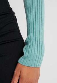 Jasnoniebieski sweter z ribbowanym rękawem wykonany z dzianiny, charakteryzujący się dopasowanym krojem. Rękaw znajduje się obok czarnej powierzchni materiału.