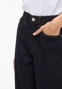 Mørkeblå denimbukser med høy midje, med oransje søm, en sølvknapp og en hånd i sidelommen.