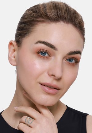 Nahaufnahme einer Person mit blauen Augen, heller Haut und neutralem Make-up; trägt einen silbernen, abstrakten Ring. Der Hintergrund ist weiß, die Haare sind zurückgebunden.