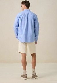 Lichtblauw overhemd met lange mouwen, met knopen bij de manchetten, gedragen met crème kleurige shorts; model staat blootsvoets en toont een neutrale achtergrond.