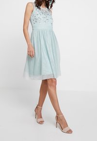 Světle modré šaty bez rukávů s vyšitými květinovými akcenty, přiléhavým korzetem a volnou sukní do kolen. Nosené s tělovými sandály na vysokém podpatku.