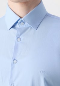 Lichtblauw overhemd met kraag, gemaakt van gladde stof, met knoopsluitingen en een geborduurd "CK"-logo op de borst.