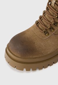 Bota de senderismo de ante marrón con punta redondeada, suela de goma beige, dibujo pronunciado y herrajes metálicos para los cordones. Área de la punta ligeramente desgastada.
