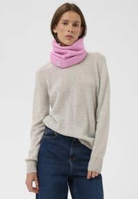 Rózsaszín kötött garbónyakú sál bordázott textúrával és címkés részlettel. Világosszürke pulóverrel és sötétkék farmernadrággal párosítva.