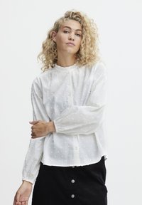 Weiße Bluse mit Rüschensaum am Ausschnitt, Knopfleiste und strukturierter Punktmusterung; lange Ärmel mit elastischen Bündchen. Kombiniert mit einem schwarzen Rock.
