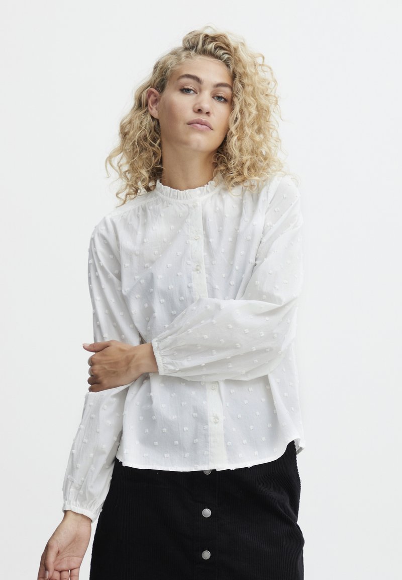 Weiße Bluse mit Rüschensaum am Ausschnitt, Knopfleiste und strukturierter Punktmusterung; lange Ärmel mit elastischen Bündchen. Kombiniert mit einem schwarzen Rock.